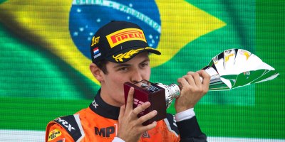 Felipe Drugovich, de macacão laranja e boné, beijando troféu de vencedor na F2 2022 com bandeira do Brasil ao fundo