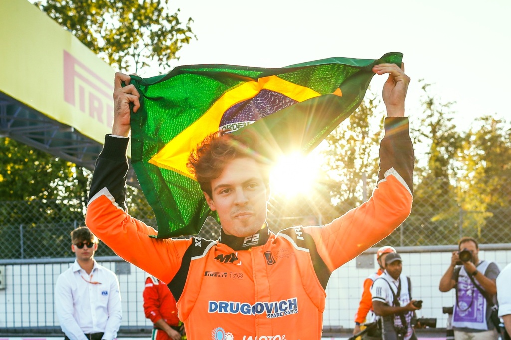 Felipe Drugovich, de macacão laranja, celebrando o título da F2 segurando a bandeira do Brasil, com o Sol ao fundo