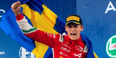 Dino Beganovic, de boné preto e uniforme vermelho cor da Ferrari, comemora o título da F-Regional by Alpine erguendo o punho e segurando a bandeira da Suécia